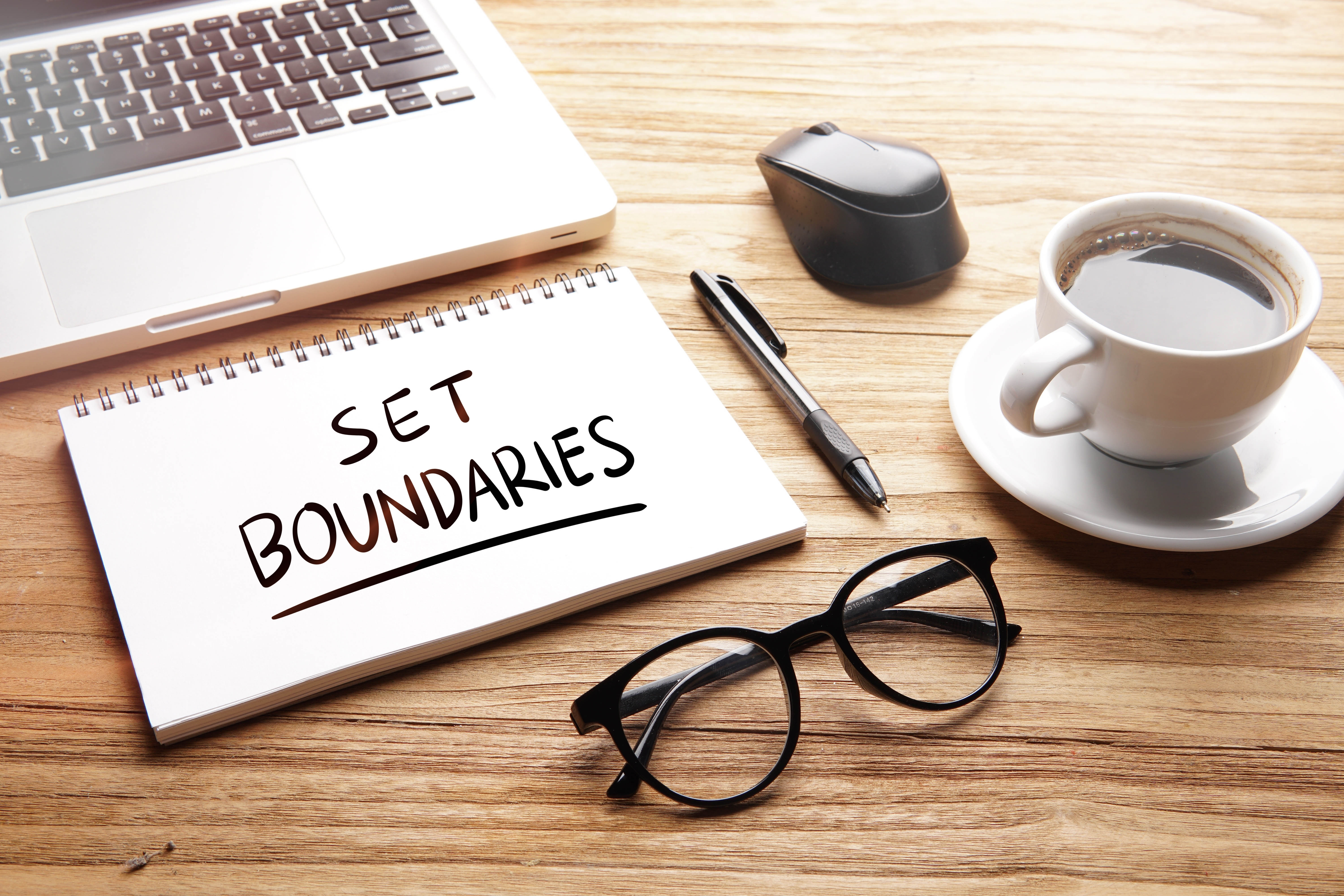 Close-up of a desk with a laptop and a notebook that says "Set boundaries" with glasses and coffee cup